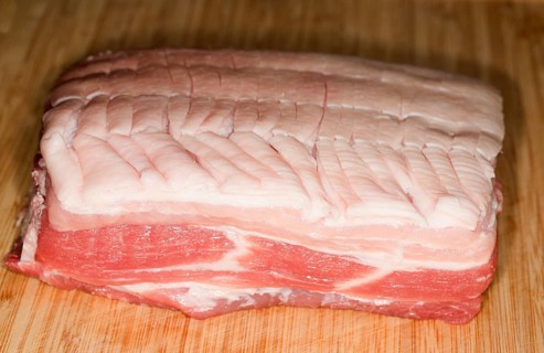 Raw lean pork belly with carved skin lies on a wooden board