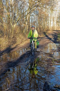 Woman cycling with e-bike over muddy forest path and through puddles