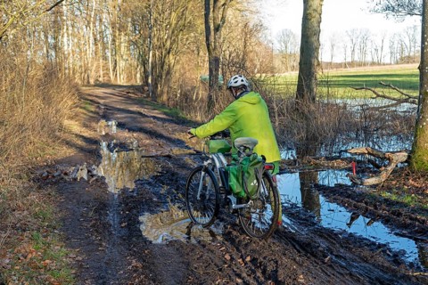 Woman cycling with e-bike over muddy forest path and through puddles