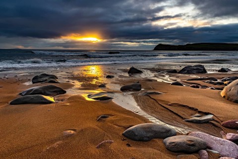 Sandy beach beach with stones on North Atlantic with rocky outcrop and cloudy sky at sunset