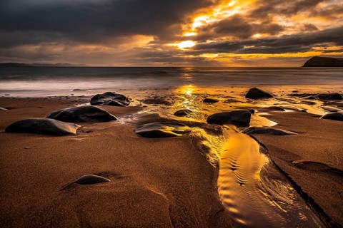 Sandy beach beach with stones on North Atlantic with rocky outcrop and cloudy sky at sunset