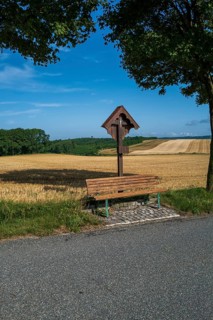 A wayside cross with a park bench at the edge of a wheat field in summer
