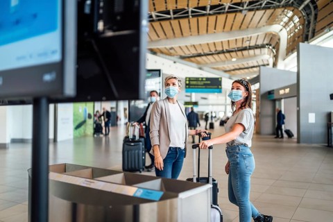 Two travelling women wearing protective masks discussing by flight information board at the Faro airport