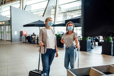 Two travelling women wearing protective masks discussing by flight information board at the Faro airport