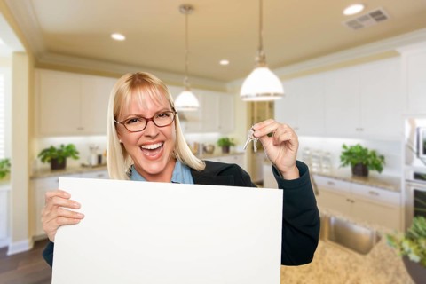 Happy young woman holding blank sign and keys inside beautiful custom kitchen