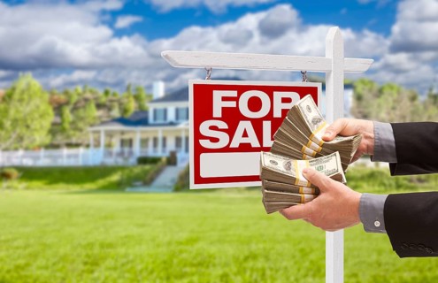 Man handing over thousands of dollars in front of house and for sale real estate sign