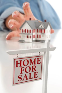 Real estate sign in front of womans hand reaching for model house on a white surface