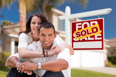 Young happy hispanic young couple in front of their new home and sold for sale real estate sign