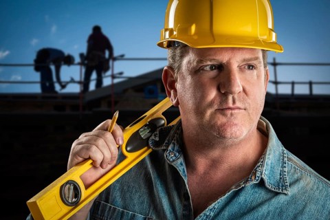 Serious contractor in hard hat holding level and pencil at construction site