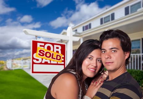 Young happy hispanic couple in front of their new home and sold for sale real estate sign