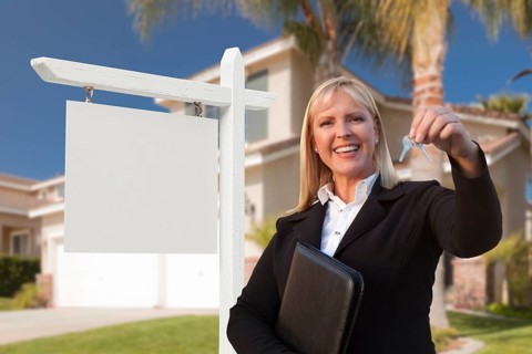Female real estate agent handing over the house keys in front of a beautiful new home and blank real estate sign