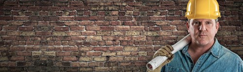 Male contractor in hard hat holding construction plans in front of old brick wall banner with copy space