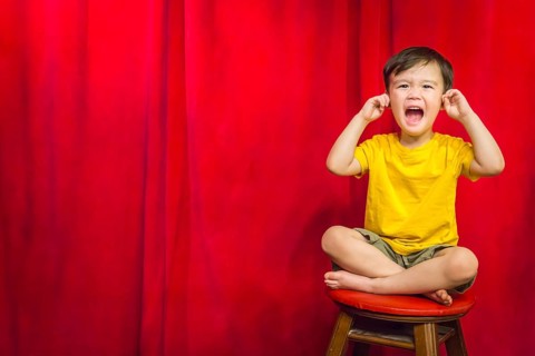 mixed-race boy with his fingers in his ears sitting on stool in front of red curtain