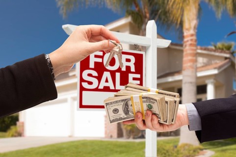 Agent handing over keys as buyer is handing over cash for house with home and for sale real estate sign behind