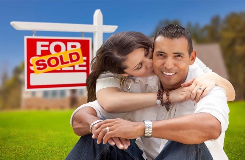 Young happy hispanic young couple in front of their new home and sold for sale real estate sign