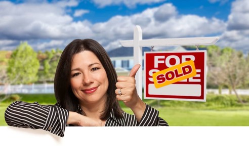 Pretty hispanic woman leaning on white with thumbs up in front of beautiful house and sold for sale real estate sign