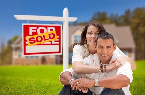 Young happy hispanic young couple in front of their new home and sold for sale real estate sign