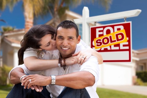Young happy hispanic young couple in front of their new home and sold for sale real estate sign