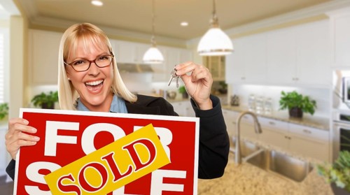 Happy young woman holding sold for sale real estate sign and keys inside beautiful custom kitchen