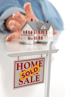 Sold real estate sign in front of womans hand reaching for model house on a white surface