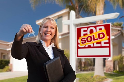 Female real estate agent handing over the house keys in front of a beautiful new home and real estate sign
