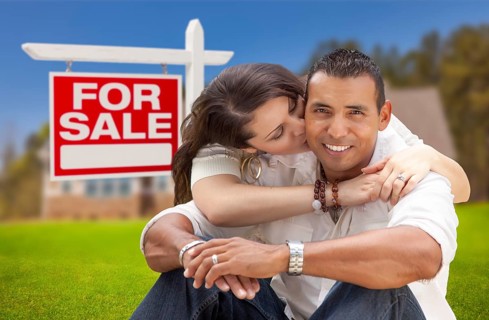 Young happy hispanic young couple in front of their new home and for sale real estate sign