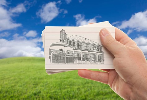 Male hand holding stack of paper with house drawing over empty grass field and sky