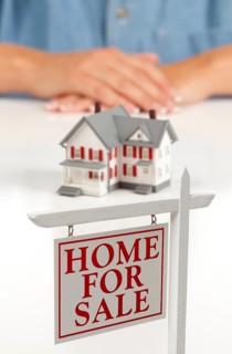 Womans folded hands behind model house and home for sale real estate sign in front on white surface