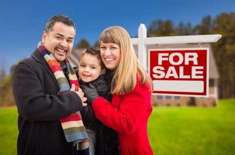 Warmly dressed young mixed-race family in front of home for sale real estate sign and house