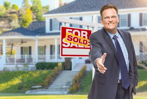 Smiling male agent reaching for a hand shake in front of beautiful house and sold real estate sign