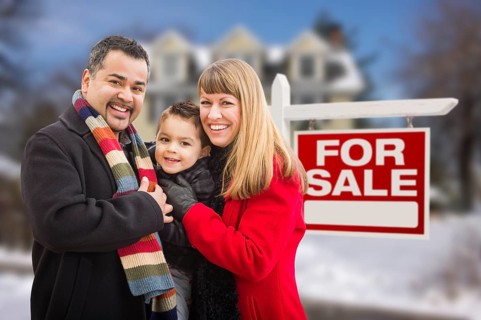 Warmly dressed young mixed-race family in front of home for sale real estate sign and house with snow on the ground