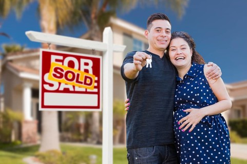 Young happy pregnant hispanic young couple with house keys in front of their new home and sold for sale real estate sign