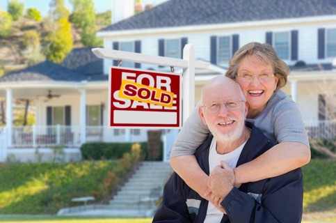 Senior adult couple in front of sold home for sale real estate sign and beautiful house