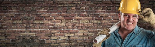 Male contractor in hard hat holding construction plans in front of old brick wall banner with copy space