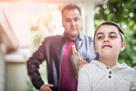 Afraid and bruised mixed-race boy in front of angry man holding bottle of alcohol