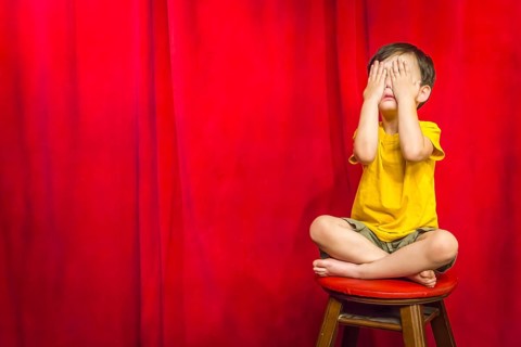 mixed-race boy boy covering his eyes sitting on stool in front of red curtain