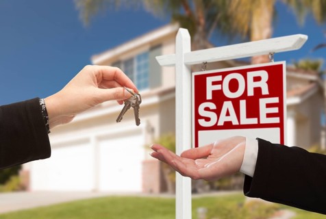 Real estate agent handing over the house keys in front of a beautiful new home and for sale real estate sign