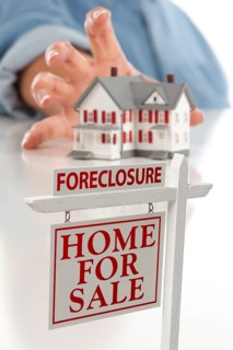 Foreclosure real estate sign in front of womans hand reaching for model house on a white surface
