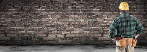 Contractor wearing hard hat and tool belt facing blank old brick wall banner with copy space