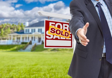 Real estate agent reaches for handshake with sold sign and new house behind