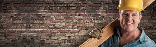 Male contractor in hard hat holding wood plank in front of old brick wall banner with copy space
