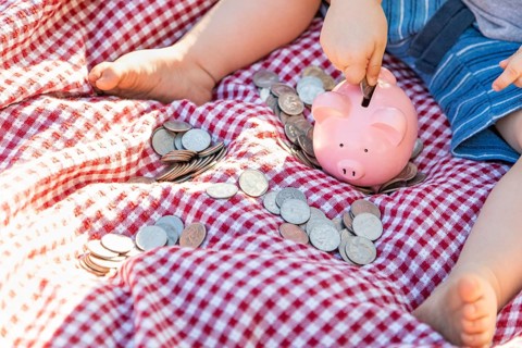 Baby boy sitting on picnic blanket putting coins in piggy bank