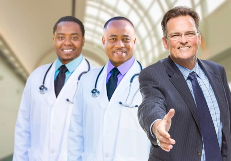 Two mixed-race doctors behind businessman reaching for a hand shake inside hospital