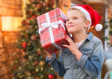 Curious young boy wearing santa hat holding christmas gift in front of decorated tree