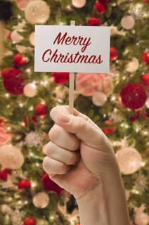 Hand holding merry christmas card in front of decorated christmas tree