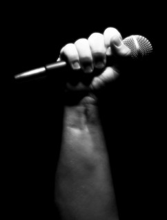 Gray scale vertical microphone clinched firmly in male fist on a black background