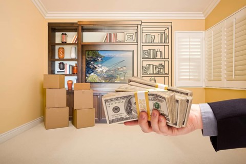 Hand holding out cash over drawing of entertainment unit in room with moving boxes