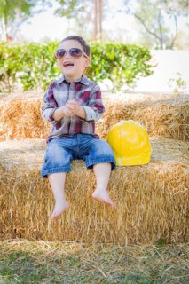 Cute young mixed-race boy laughing with sunglasses and hard hat outside sitting on hay bale