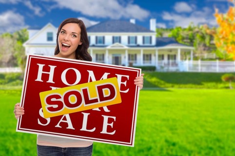 Excited mixed-race female with sold home for sale real estate sign in front of beautiful house