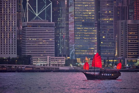 Tourist junk boat ferry with red sails and Hong Kong skyline cityscape downtown skyscrapers over Victoria Harbour in the evening. Hong Kong